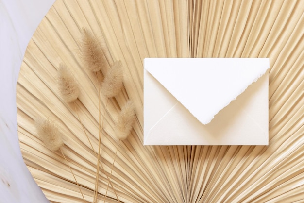 Square envelope on dry beige palm leaf near hare's tail grass top view boho wedding mockup