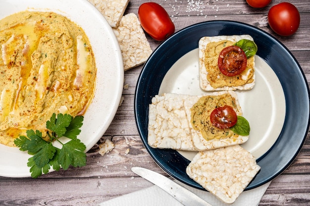 Square rice crackers with hummus and cherry tomatoes on a plate Tomatoes crackers on a rustic table and a platter with hummus Natural and healthy food concept