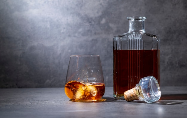 Square whiskey bottle and glass with ice on dark gray cement surface