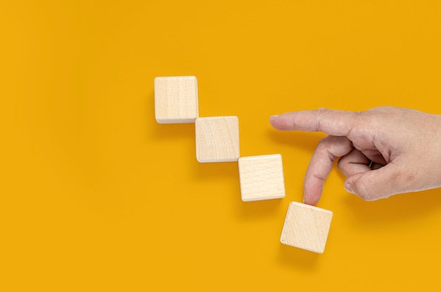 Square wooden blocks are placed on a yellow background, hands are walking upwards with the last piece seeming to collapse. Wood block concept, banner with copy space for text, poster, mockup template.