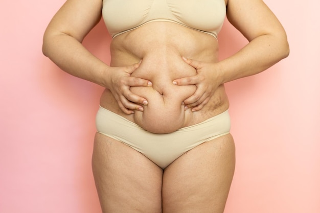 Squeeze woman sagging belly closeup folds on tummy loose skin and cellulite obesity Naked overweight plus size girl on pink background in beige underwear Concept of dieting and body control