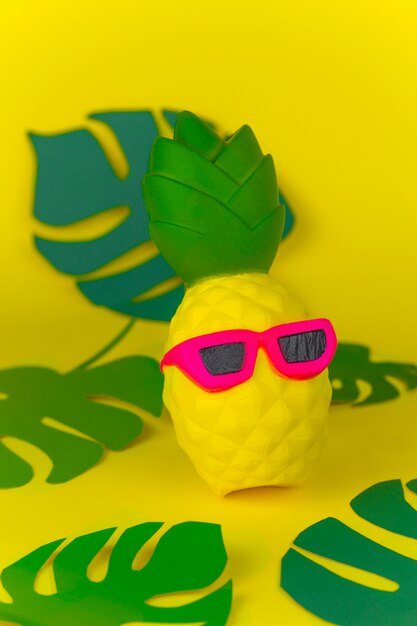 Squishy toy pineapple in sunglasses on yellow background among paper cut tropical leaves