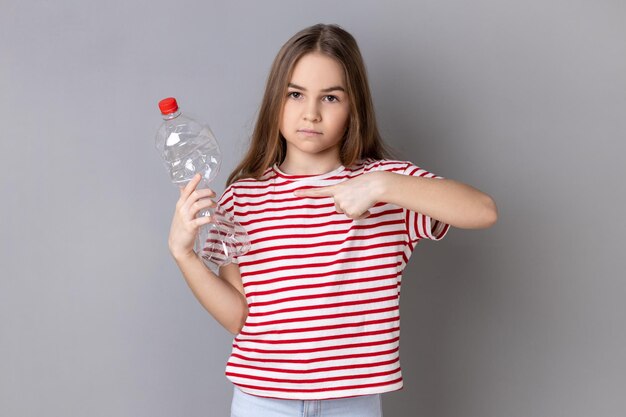 Srious charming little girl in striped Tshirt holding and pointing finger at empty plastic bottle