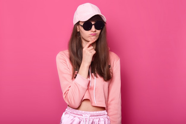 Srusio shot of stylish teenager girl wearing fashionable shortened pink hoodie, cap and black subglasses, touches her chin with thoun ghtful facialexpression. female. People concept.