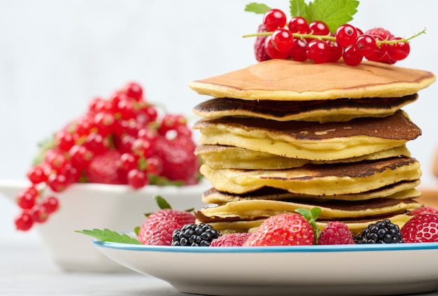 Stack of baked pancakes with fruits in a round plate on a white table