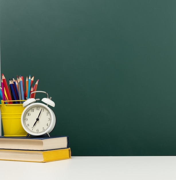 Stack of books, a round alarm clock and multicolored pencils on the background of an empty green chalk board. Back to school