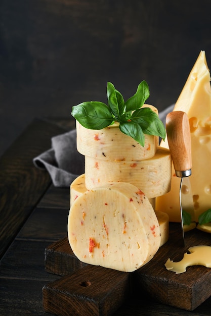 Stack of cheese with tomatoes and seasoning and Swiss cheese on with basil and knife on serving board on dark wooden table background Assortment of different cheese types Cheese background