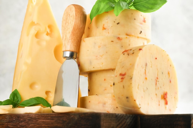 Stack of cheese with tomatoes and seasoning and Swiss cheese on with basil and knife on serving board on light grey table background Assortment of different cheese types Cheese background