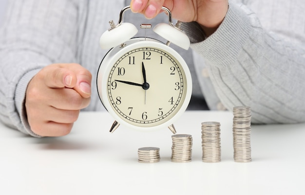 Stack of coins and round white alarm clock on the table. The concept of time is money, a period of discounts.