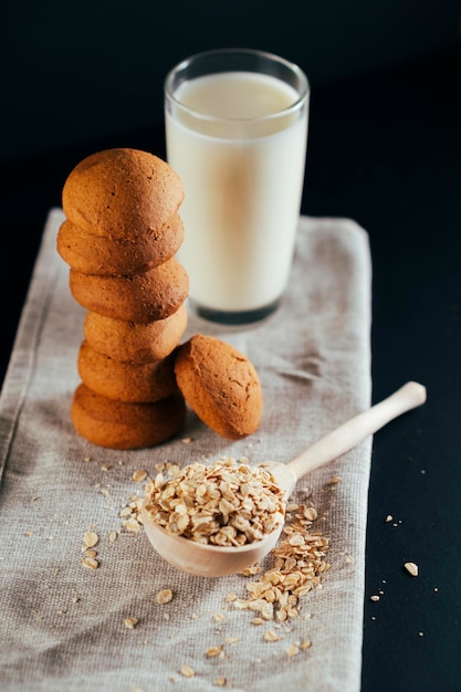 Stack of fresh oatmeal cookies with milk in glass raw oat flakes in large wooden spoon on black background Stilllife