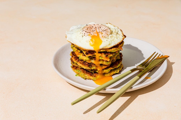 A stack of fried vegetable pancakes made from zucchini and carrot with a fried egg with runny yolk on top shot with hard light with shadows served on a plate with golden knife and fork