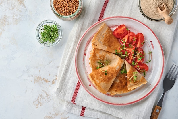 Stack of gluten free buckwheat flour crepes pancakes with cherry tomatoes and arugula microgreens on white plate, homemade healthy baking for breakfast. Mock up. Copy space.