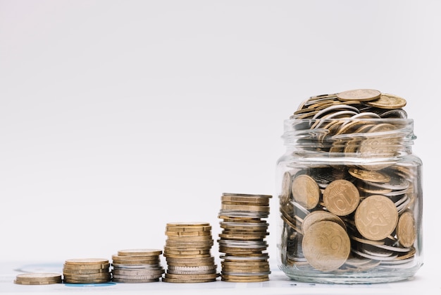 Stack of increasing coins with jar filled with coins against white backdrop