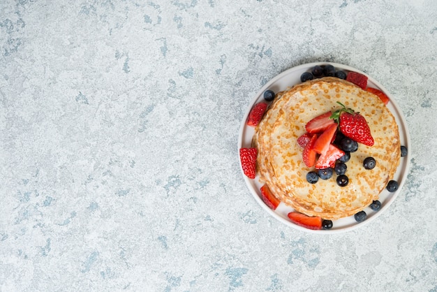 A stack of thin homemade pancakes lies in a plate , decorated with berries and honey, on a gray concrete background, close- up, copy space