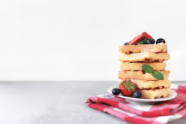 Stack of waffles on a white plate on the towel and table with blueberry, chopped strawberry and mint leaves. High quality photo