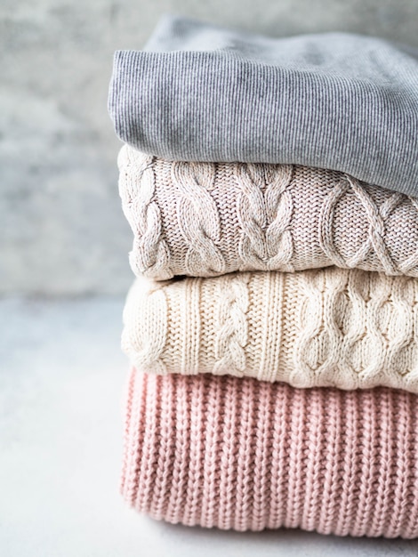 Stack of warm knitted woolen sweaters pastel shades against grey wall.