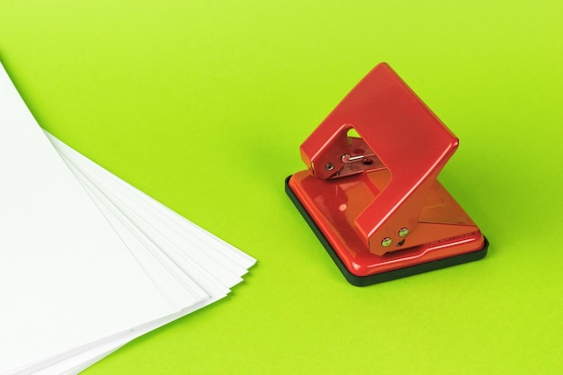 A stack of white sheets of paper and a red hole punch on a green background