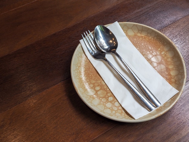 Stainless steel spoon and fork on white clean napkin on vintage style circle ceramic plate on wooden table