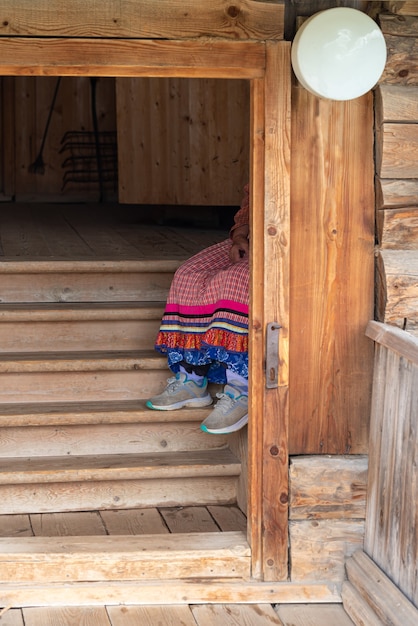 On the stairs of the hut sits a grandmother dressed in Russian folk style and modern sneakers