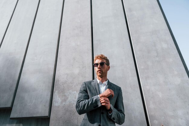 Photo standing against building wall young businessman in grey formal wear is outdoors in the city