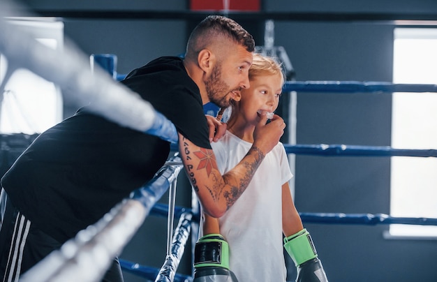 Photo standing on the boxing ring. young tattooed boxing coach teaches cute little girl in the gym.