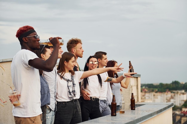 Standing high with alcohol Cloudy sky Group of young people in casual clothes have a party at rooftop together at daytime