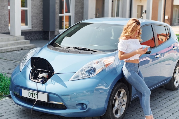 Standing near the car Young woman in casual clothes with her electromobile outdoors at daytime