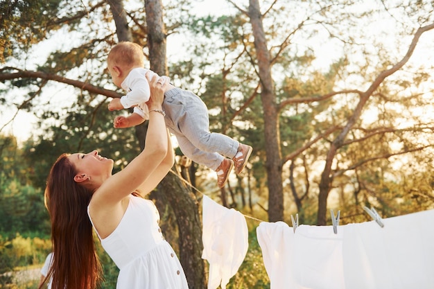 Standing near white clothes that hanging on the rope to dry Young mother with her little son is outdoors in the forest Beautiful sunshine