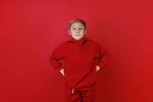 Standing on a red background A boy dressed in a red tracksuit He holds his hands on his belt