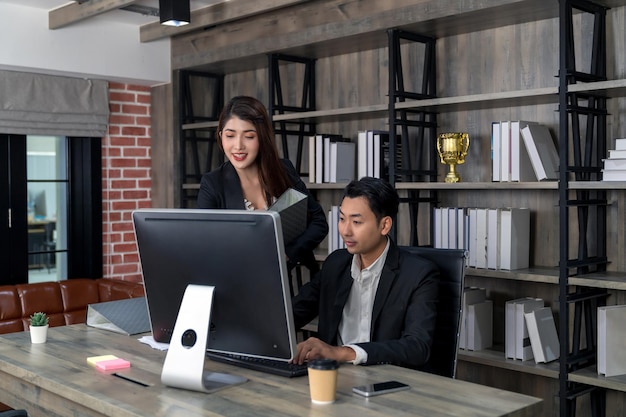 Standing smart businesswoman present data analysis on computer monitor to sitting businessman in modern wooden office happy Asian business people working together looking at laptop