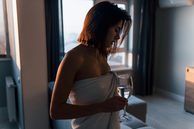 Standing at sunrise time in white towel Young woman is indoors in room of modern house at daytime