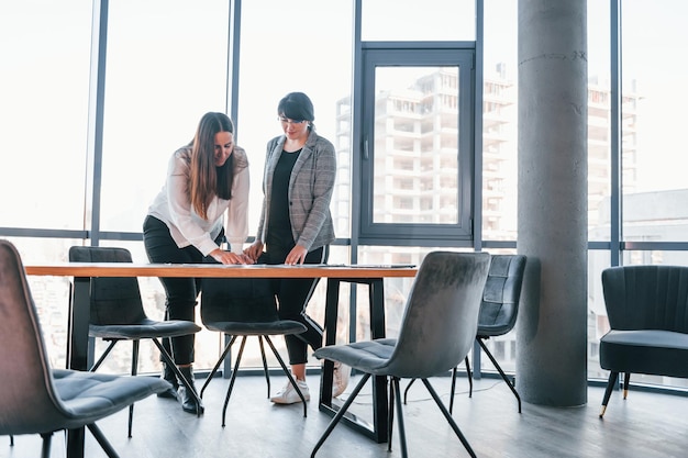 Standing and talking Two women in formal clothes is indoors in the modern office works together