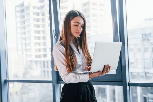 Standing with laptop Young woman in white formal clothes is indoors in the modern office