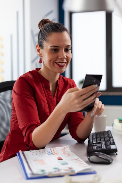 Startup manager in videocall conference using smartphone. Entrepreneur using mobile phone in startup business office. Smiling buinesswoman in red shirt browsing internet in front of desktop computer.