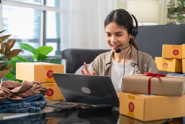 Startup small business concept woman owner wearing microphone headset of customer support phone operator and packing on the box to customer at home office seller prepares the delivery