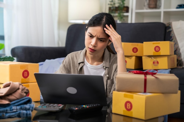 Startup small business concept, young woman owner hands touch on the forehead have a headache because of stress, and checking online order on digital laptop with packing on the box at home office.