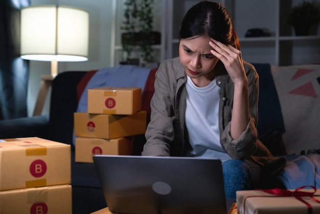 Startup small business concept young woman owner hands touch on the forehead have a headache because of stress and checking online order on digital laptop with packing on the box at home office