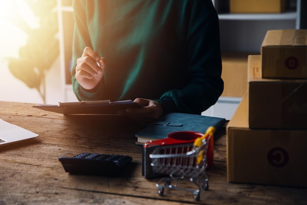 Startup SME small business entrepreneur of freelance Asian woman wearing apron using laptop and box to receive and review orders online to prepare to pack sell to customers online sme business ideas