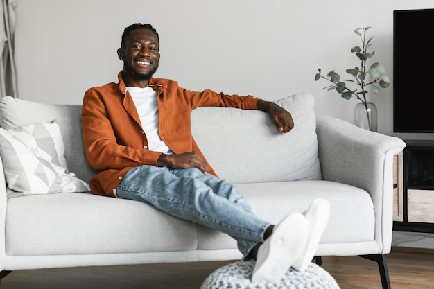 Stay at home alone enjoy silence Happy african american man sitting on sofa in cozy light living room copy space