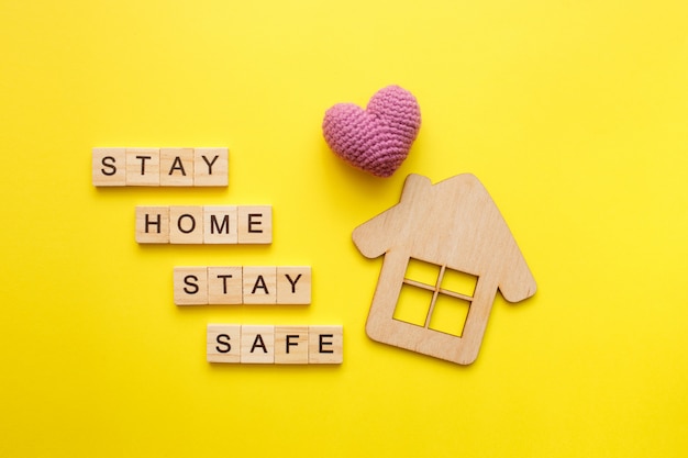 Stay Home text with wooden house with knitted heart on yellow background. Quarantine concept, covid-19 virus protection. Top view, flat lay.