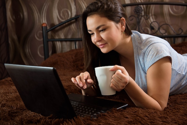 Stay home. Work home. Brunette girl using laptop. Young woman in casual clothes lying on the bed at cozy home interior.