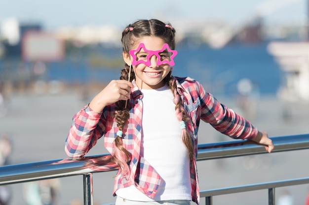 Staying focused with glasses. Small kid smiling with fancy party glasses on urban background. Funny girl holding star shaped glasses on stick. Little child looking happy through fashion prop glasses.