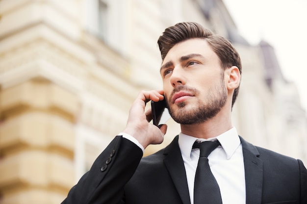 Staying in touch. Low angle view of confident young man in formalwear holding mobile phone and looking away while standing outdoors