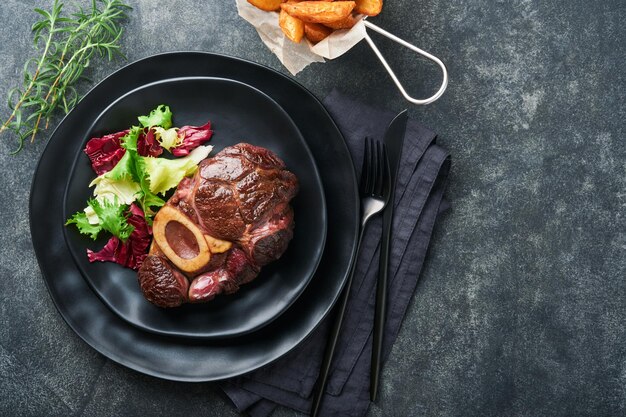 Steaks Osso Buco Sliced beef grilled Osso Buco or porterhouse meat steak with garnished with salad and french fries on black marble board on old wooden background Top view Mock up