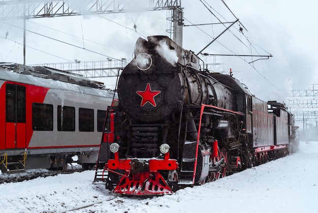 Steam locomotive and modern electric multiple-unit train stand nearby at station in winter