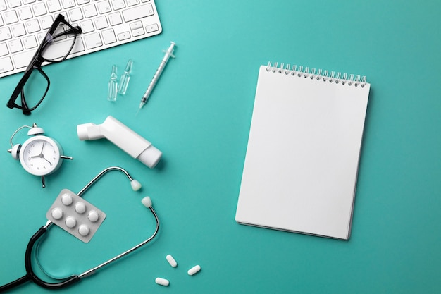 Stethoscope in doctors desk with keyboard, alarm clock, glasses, inhaler, syringe, ampoules, inhaler and pills. Top view with place for your text.