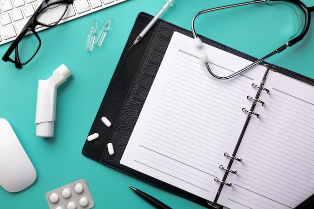 Stethoscope in doctors desk with notebook, keyboard, mouse, glasses, syringe, ampoules, inhaler and pills. Top view with place for your text.