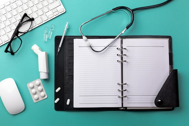 Stethoscope in doctors desk with notebook, keyboard, mouse, glasses, syringe, ampoules, inhaler and pills. Top view with place for your text.