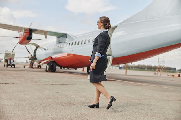 Stewardess walking on runway near airplane jet. Modern passenger plane. Side view of young woman wear uniform and glasses. Civil commercial aviation. Air travel concept