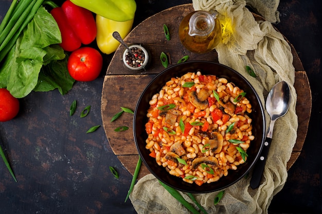 Stewed white beans with mushrooms and tomatoes with spicy sauce in a black bowl. Top view. Flat lay